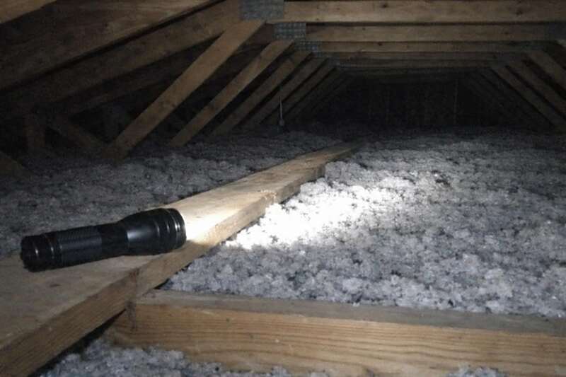 A flashlight lying on an attic joist, throwing light across the blown-in insulation and trusses