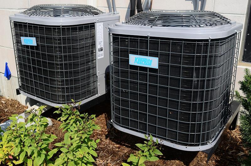 Two outdoor air conditioning condensers standing on a pad with planting around them